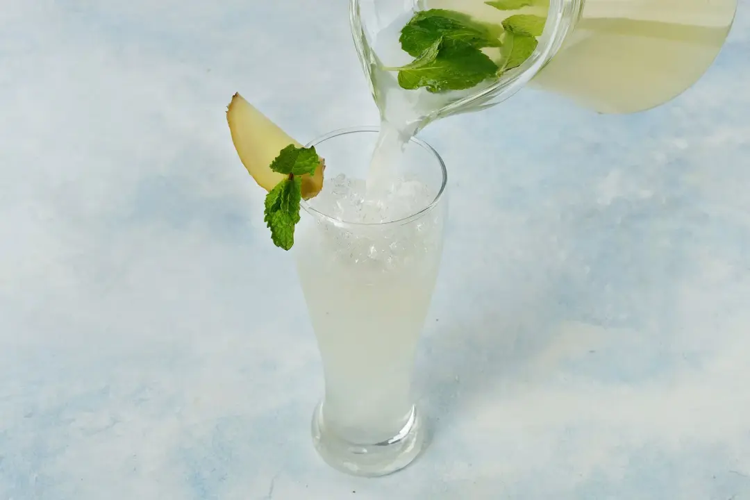 A pitcher pouring a ginger lemonade drink filled with mint leaves into a tall iced glass garnished with ginger slice and mint leaves