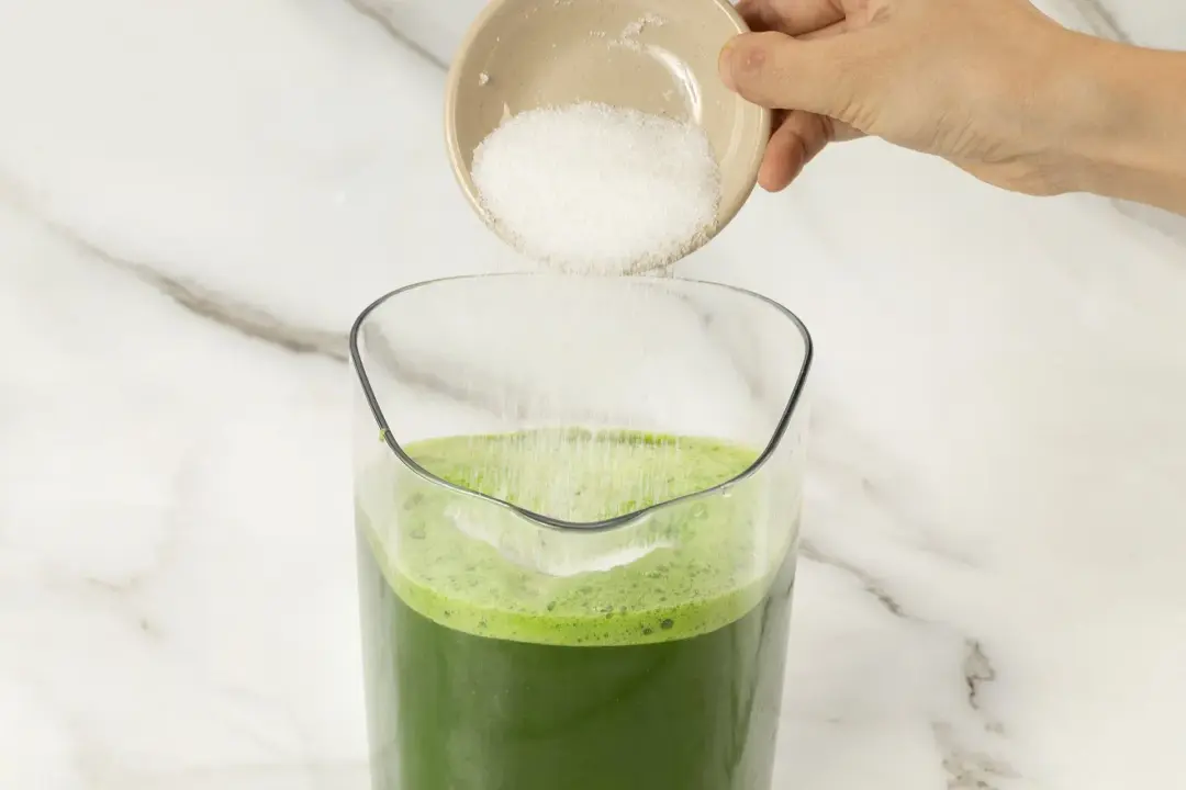 A pitcher of green machine juice with some sugar being added in