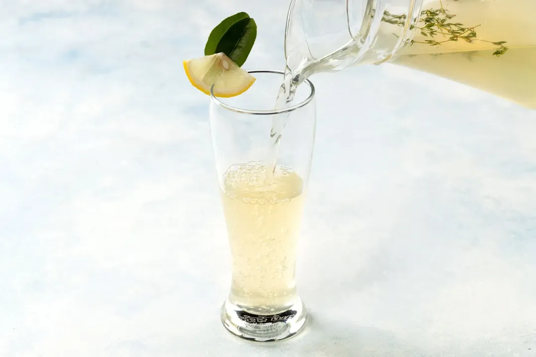 A glass of lemonade being poured from a pitcher, garnished with lemon leaves and lemon wedge