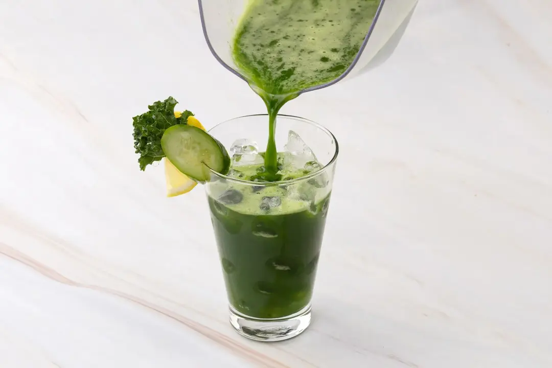 A green juice poured from a pitcher into an iced glass garnished with cucumber, kale, and pineapple