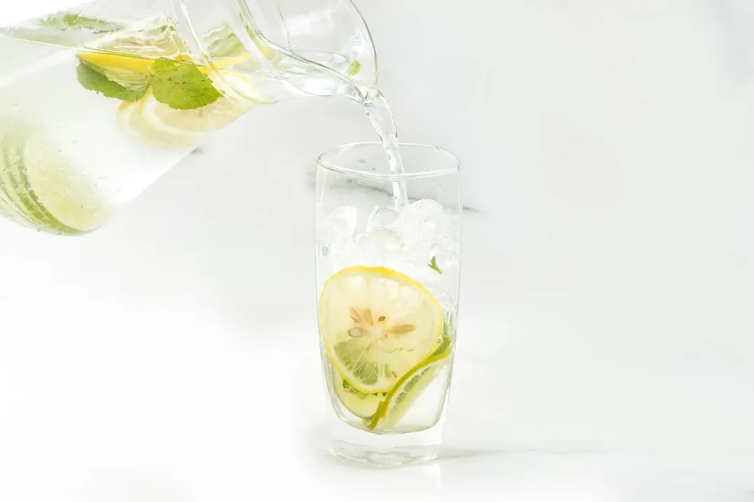 Pouring some lemon water from a pitcher into an iced glass with some lemon wheels in it