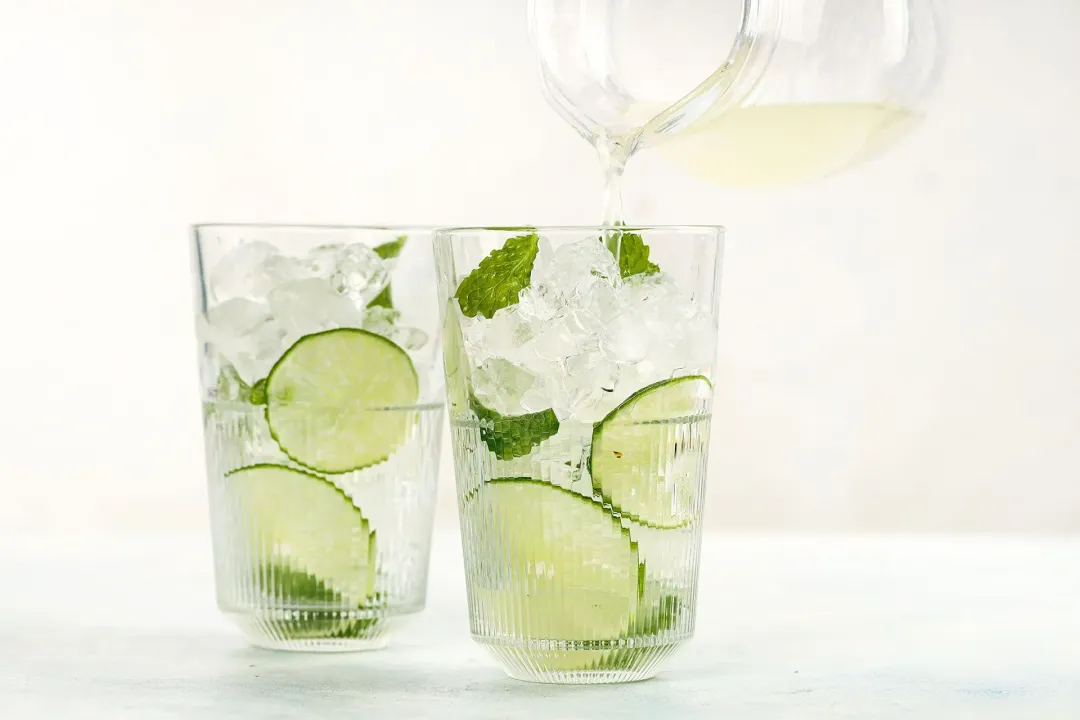 Pouring lemon juice from a pitcher into two iced glasses with lime wheels and mints inside