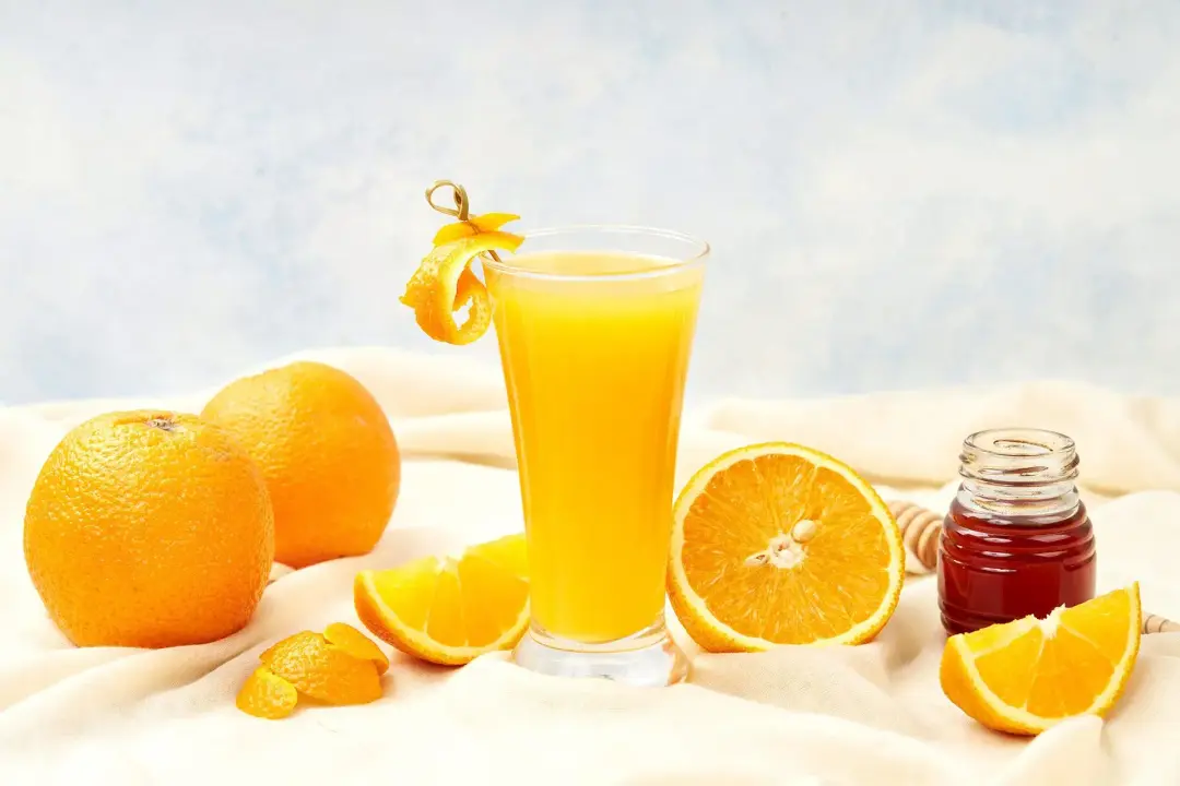 A glass of orange juice on a cloth with two oranges, half an orange, orange wedges, and a honey jar