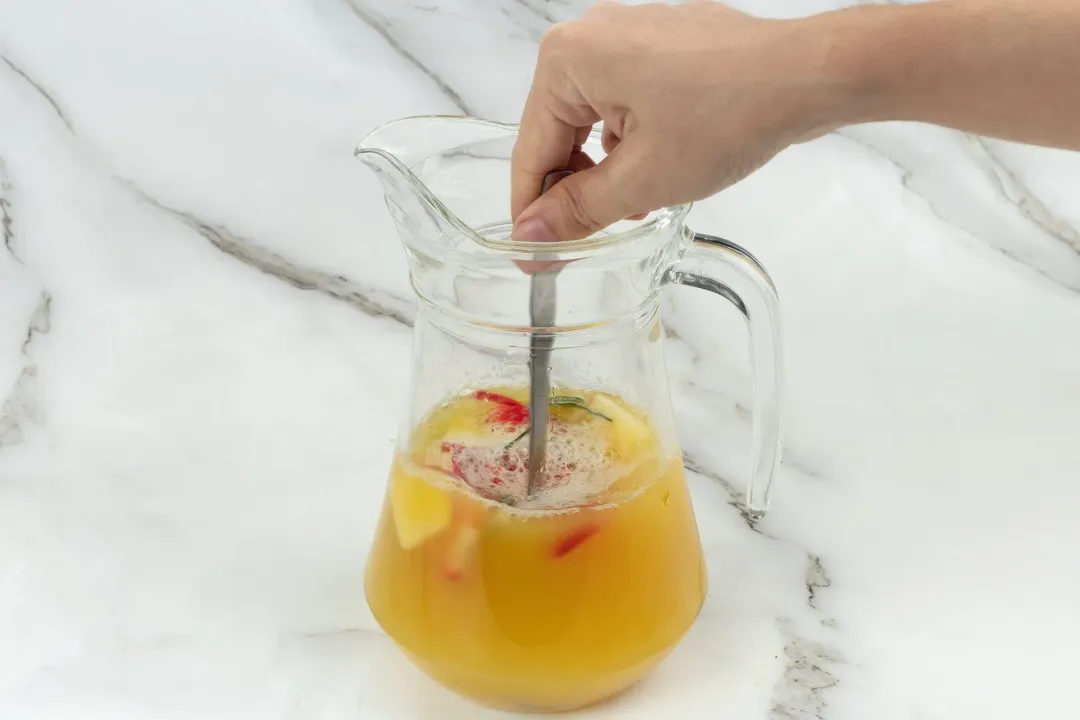 A pitcher of orange cocktail and fruits stirred with a silver spoon