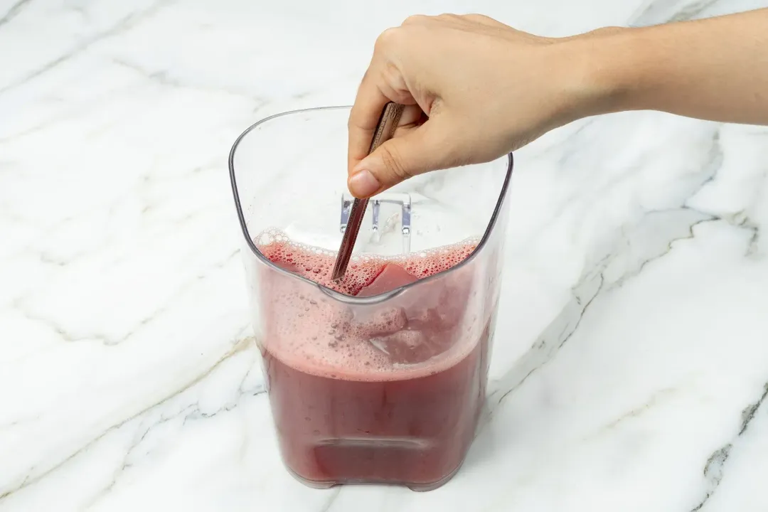 A hand holding a spoon stirring a juice container containing a burgundy beverage