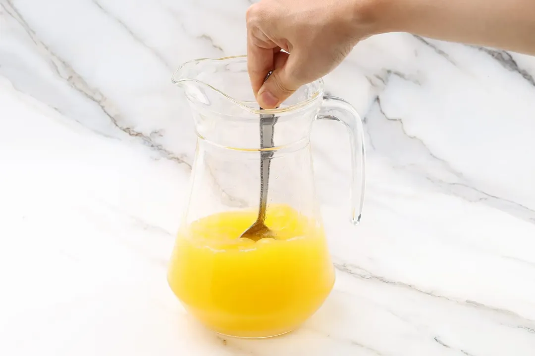 A hand dunking a spoon into a pitcher of orange juice