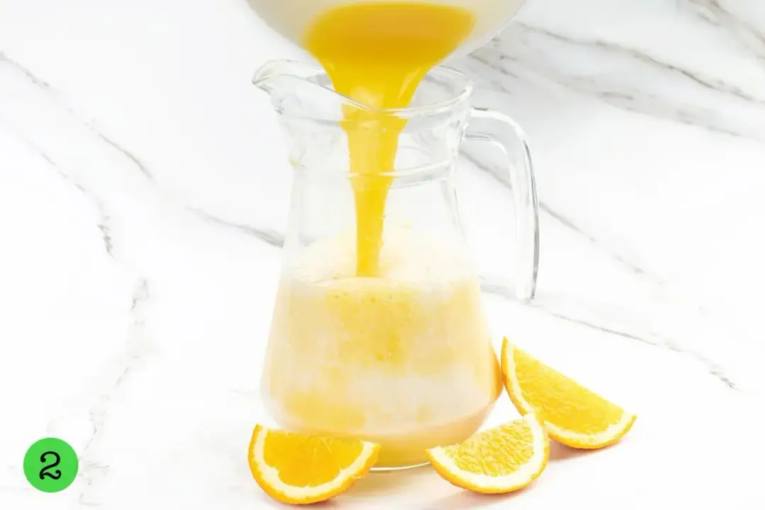 Orange juice poured into a clear glass pitcher next to some orange wedges