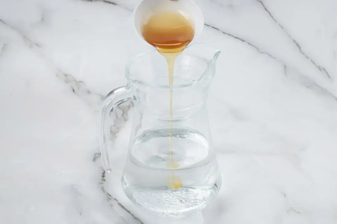 Some honey poured from a white bowl into a clear pitcher holding water