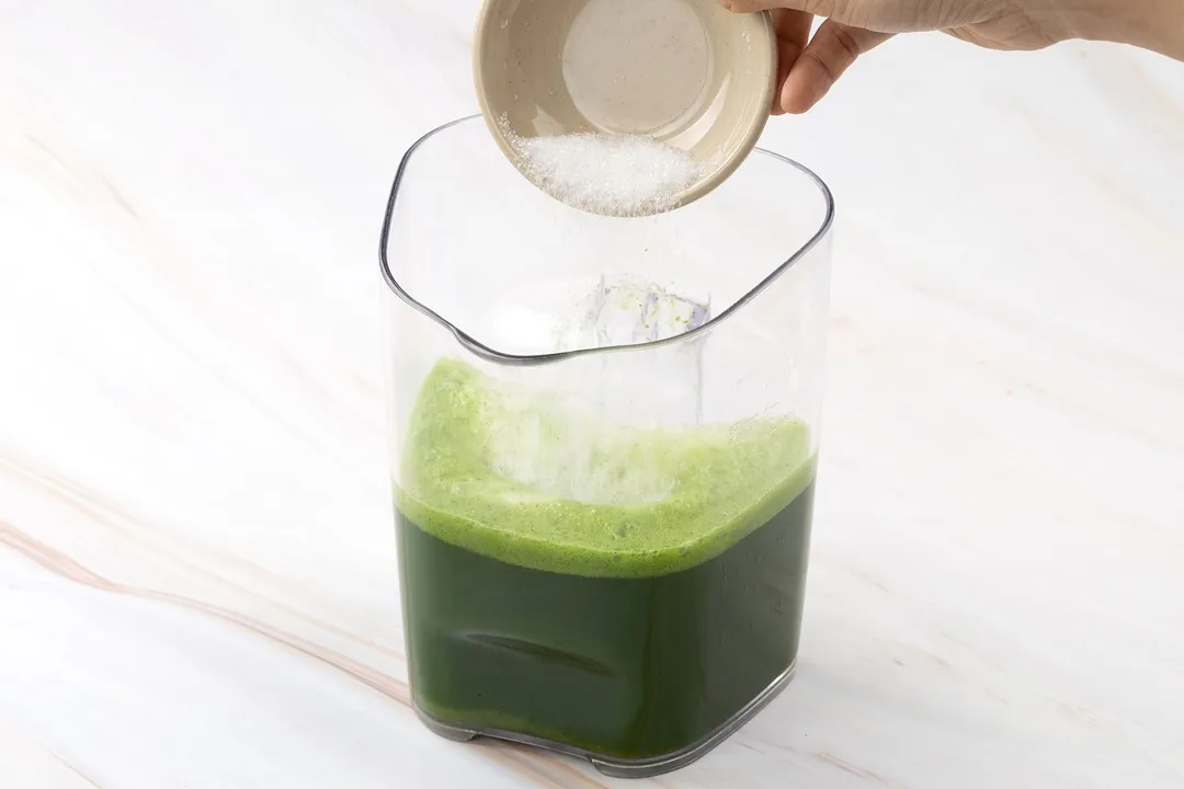 pouring sugar from a small bowl to a pitcher of green juice