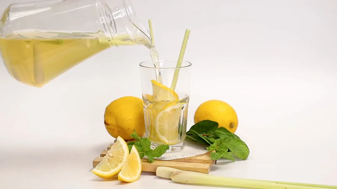 A glass jar pouring mixture of apple cider vinegar and lemon juice, with slices of lemon sliced of lemon and lemongrass in to a glass decorated with lemonm 