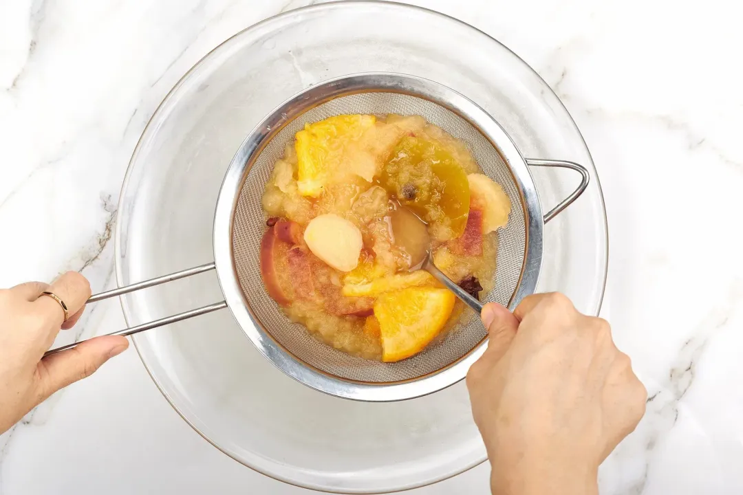 Lots of cooked fruit pulps pressed by a spoon in a sieve over a clear bowl of cider