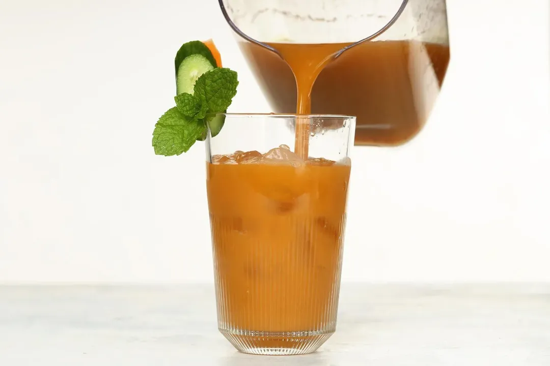 A brown drink poured from a pitcher into a clear glass with ice inside, and cucumber and mints on top