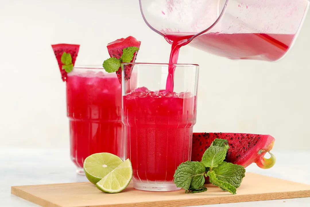 A glass jug pouring dragon fruit juice into a glass laid on a wooden board with lime wedges and mint leaves.