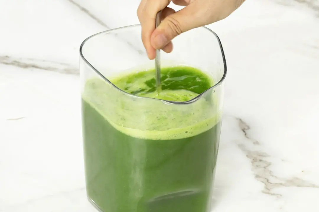 A pitcher of green machine juice being stir by a metal spoon to dissolve the added sugar