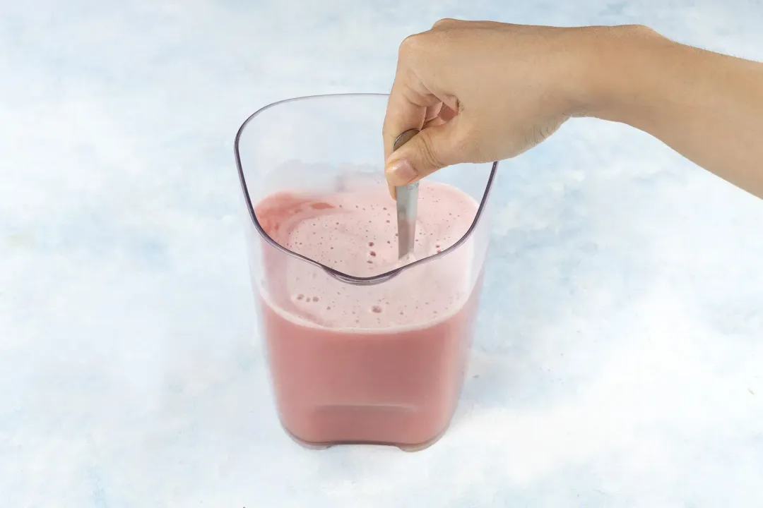 A hand holding a spoon to stir guava juice in a large juice container