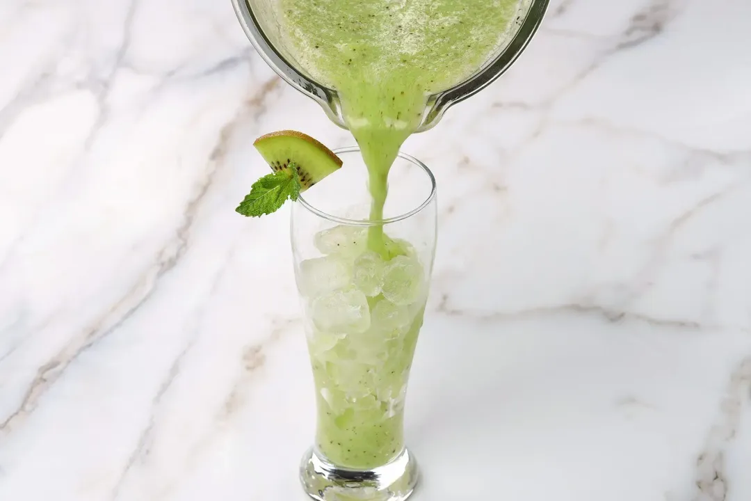 A blender pouring a green kiwi drink into an iced glass garnished with a mint leaf and a kiwi triangle