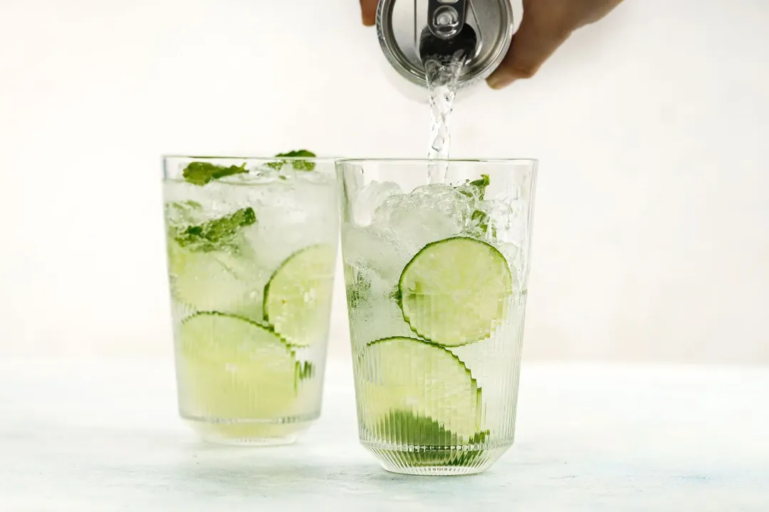 Pouring soda straight from a can into two glasses of ice, lime wheels, mints, and lime juice inside