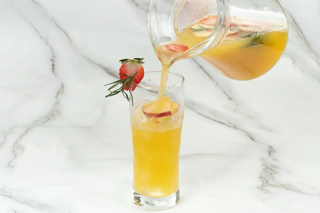 A pitcher containing an orange vodka cocktail pouring into an iced glass with a strawberry slice on the rim