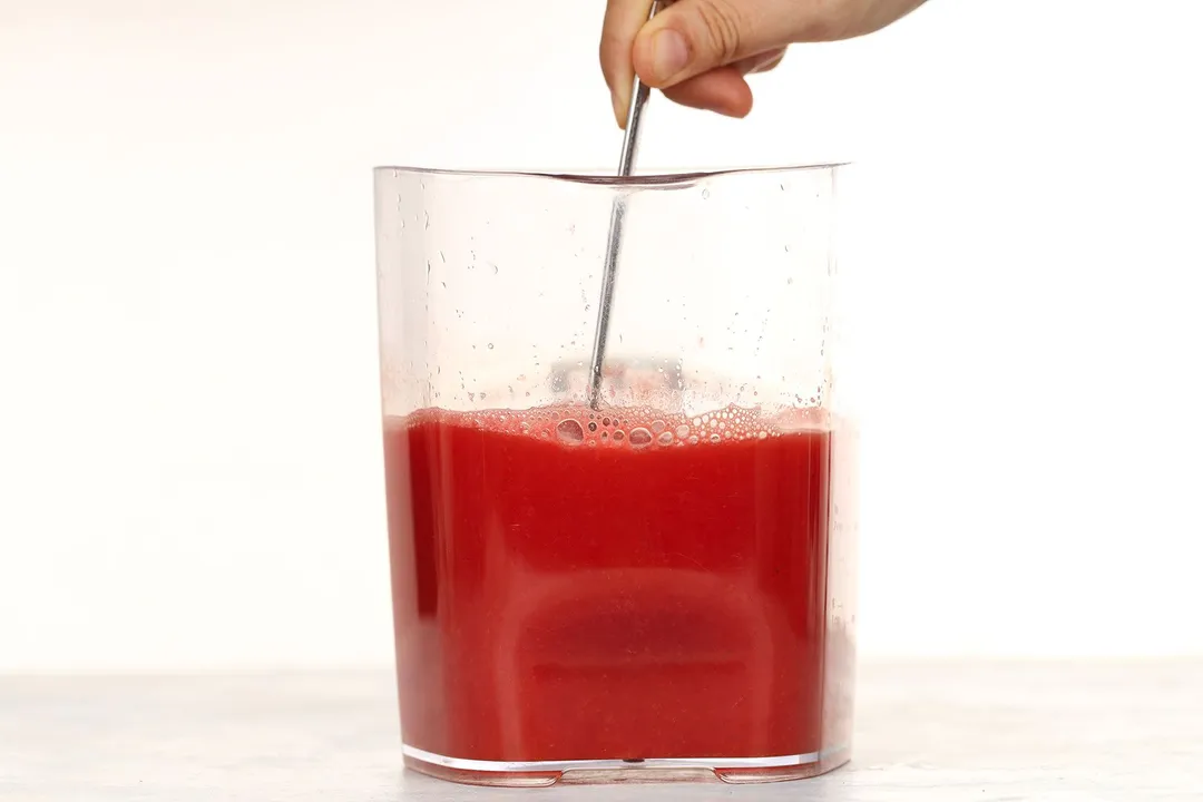 A hand using a spoon to stir in a large glass jug filled up to two-thirds with watermelon carrot juice.
