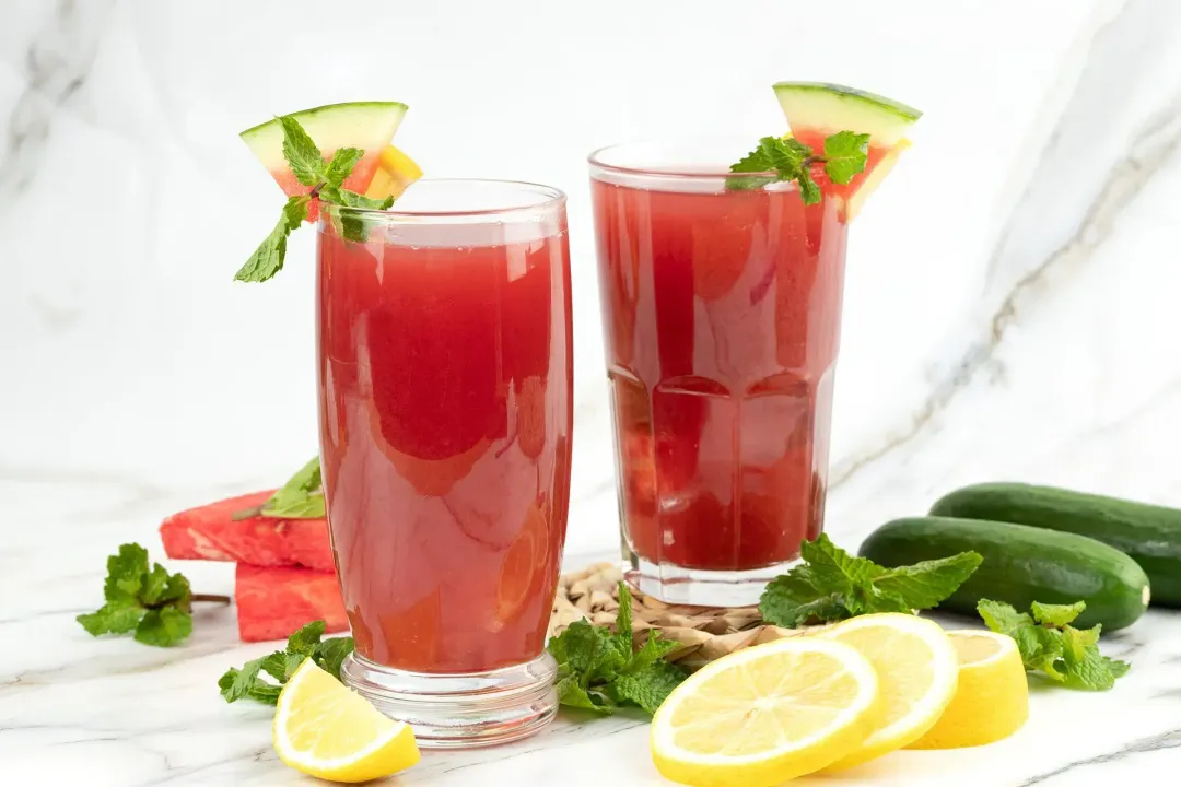 Two glasses of watermelon cucumber juice surrounded by lemon slices, spearmint leaves, watermelon slices, and whole cucumbers