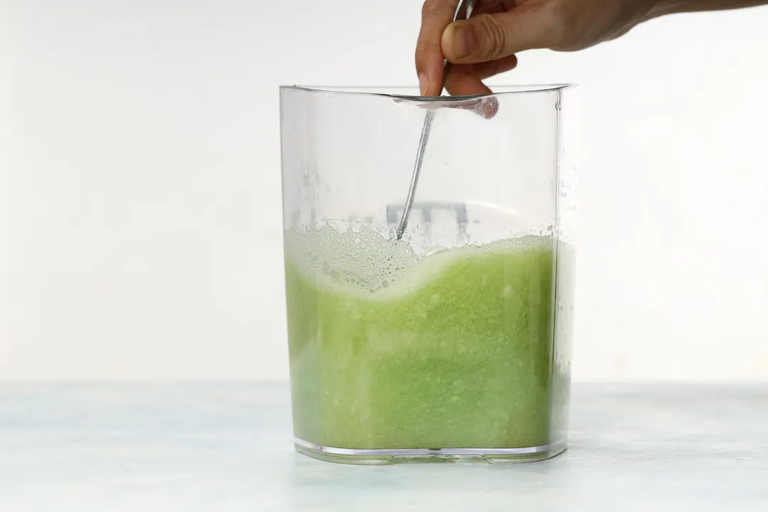 A hand using a spoon to stir in a large glass jug half filled with winter melon juice.