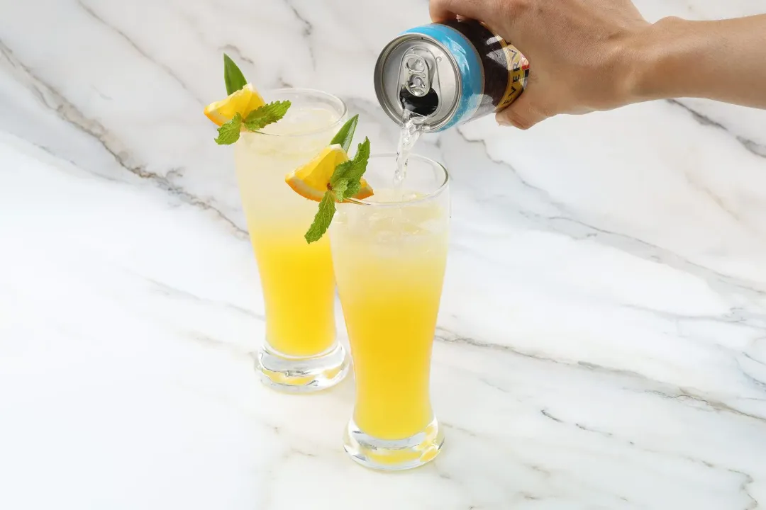 A hand pouring club soda on top of an iced orange juice glass, next to another glass of unstirred soda and orange juice