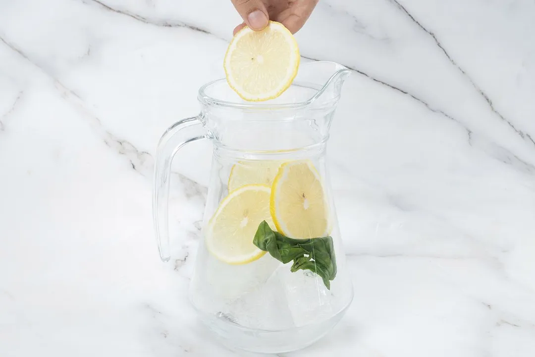 a hand holding a lemon slice on top of a jar with lemon slices and basil leaves inside