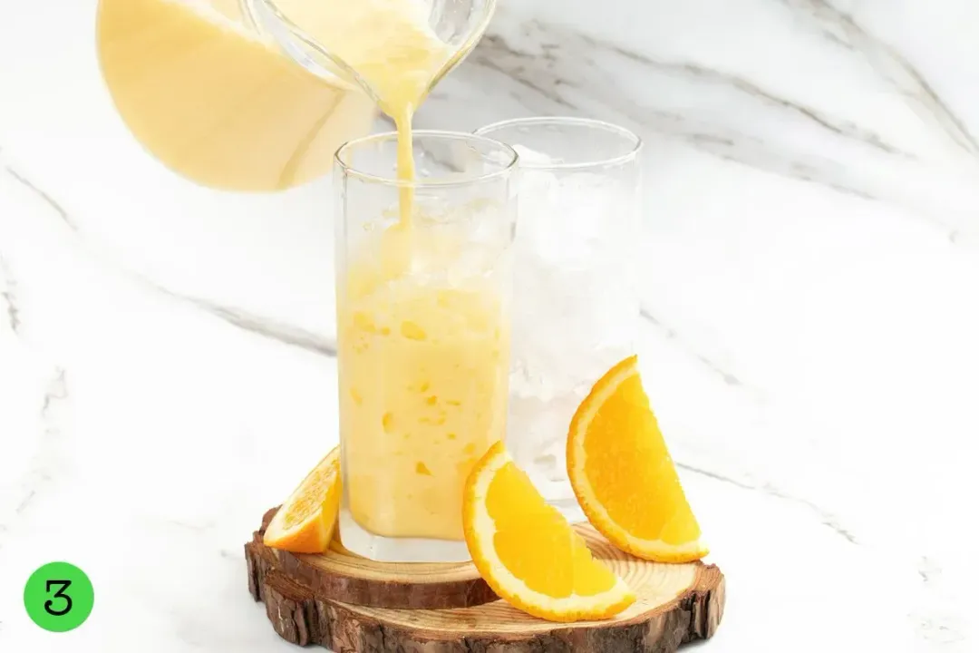 A pitcher of milk and orange juice drink poured into a tall iced glass with orange wedges around