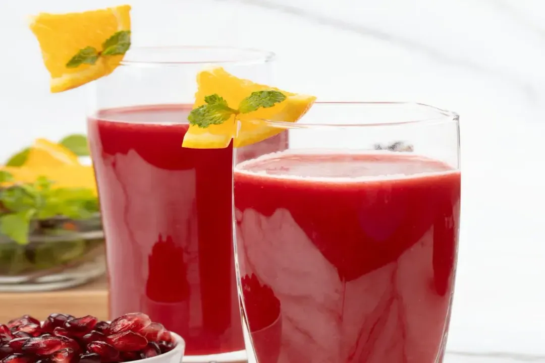 Two glasses of pomegranate orange juice put side by side, with orange wedges and mints on the rims