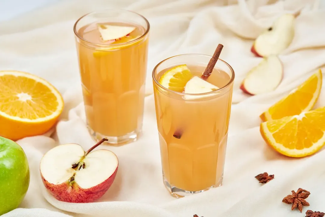 Two glasses of homemade apple cider put on a linen cloth along with apple wedges, orange wedges, and star anise
