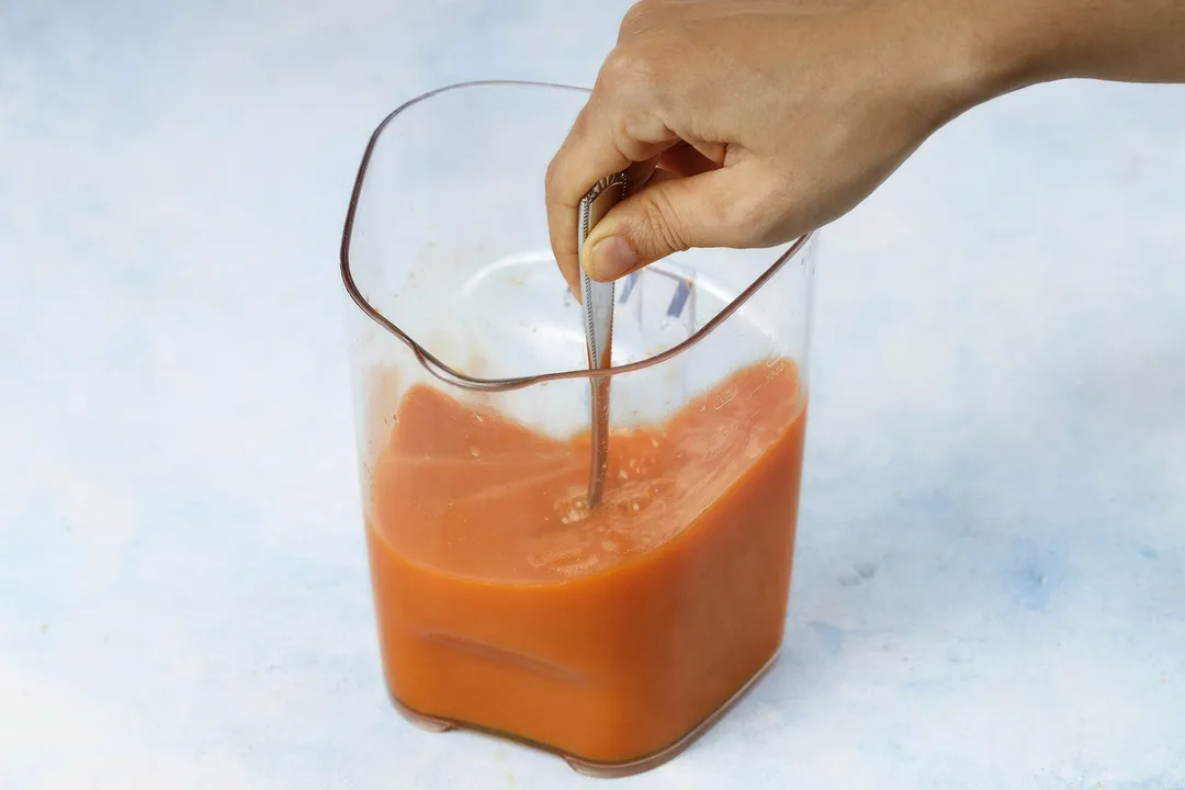 A hand using a spoon to stir Carrot Ginger Turmeric Juice in a glass jug.