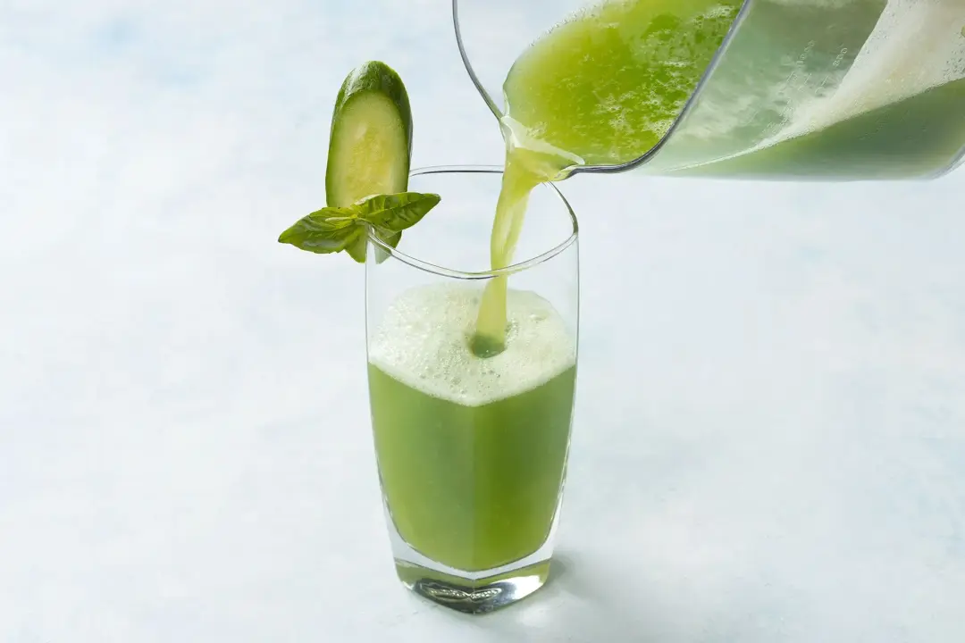 A pitcher pouring a green cucumber drink into a tall glass already garnished with a cucumber slice and mint