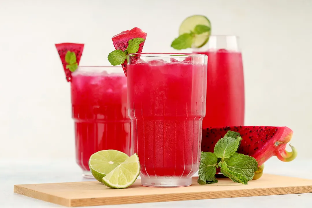 Three glasses of dragon fruit juice placed on a wooden board with a dragon fruit wedge, lime wedges, and mint leaves.