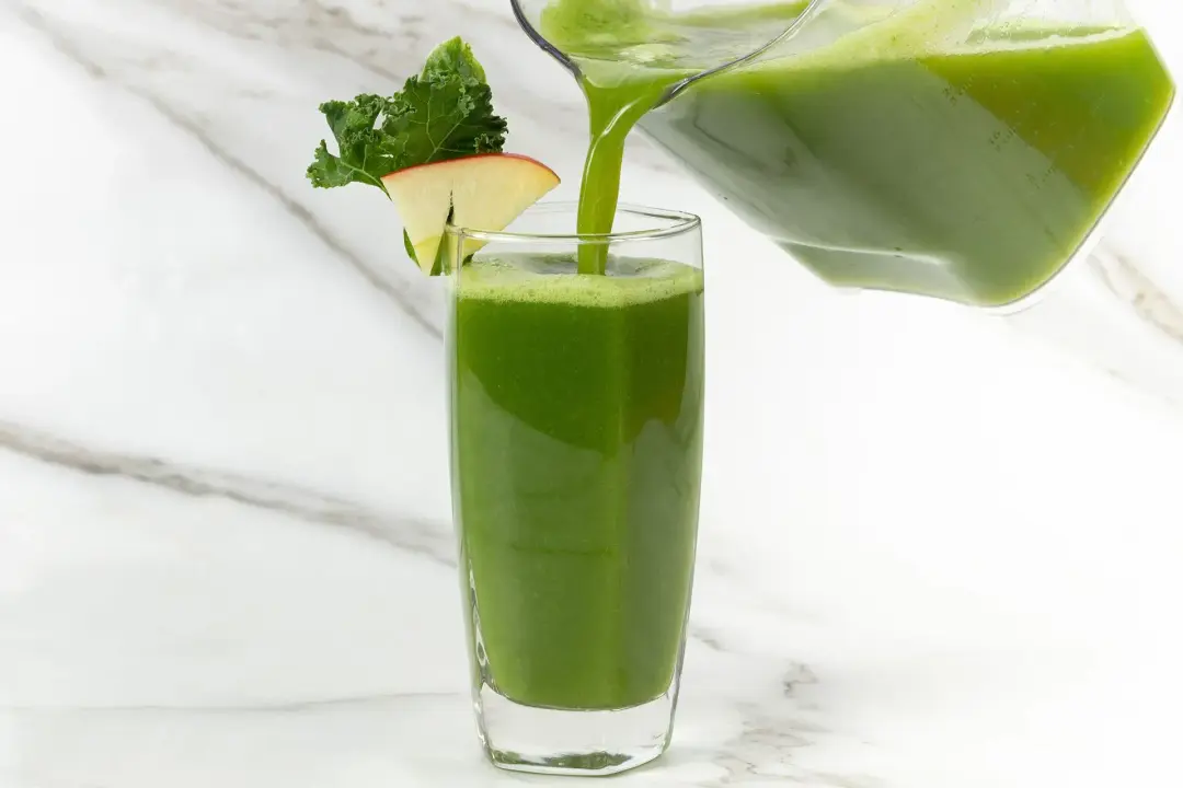 A large pitcher pouring green juice into a tall glass, garnished with an apple slice