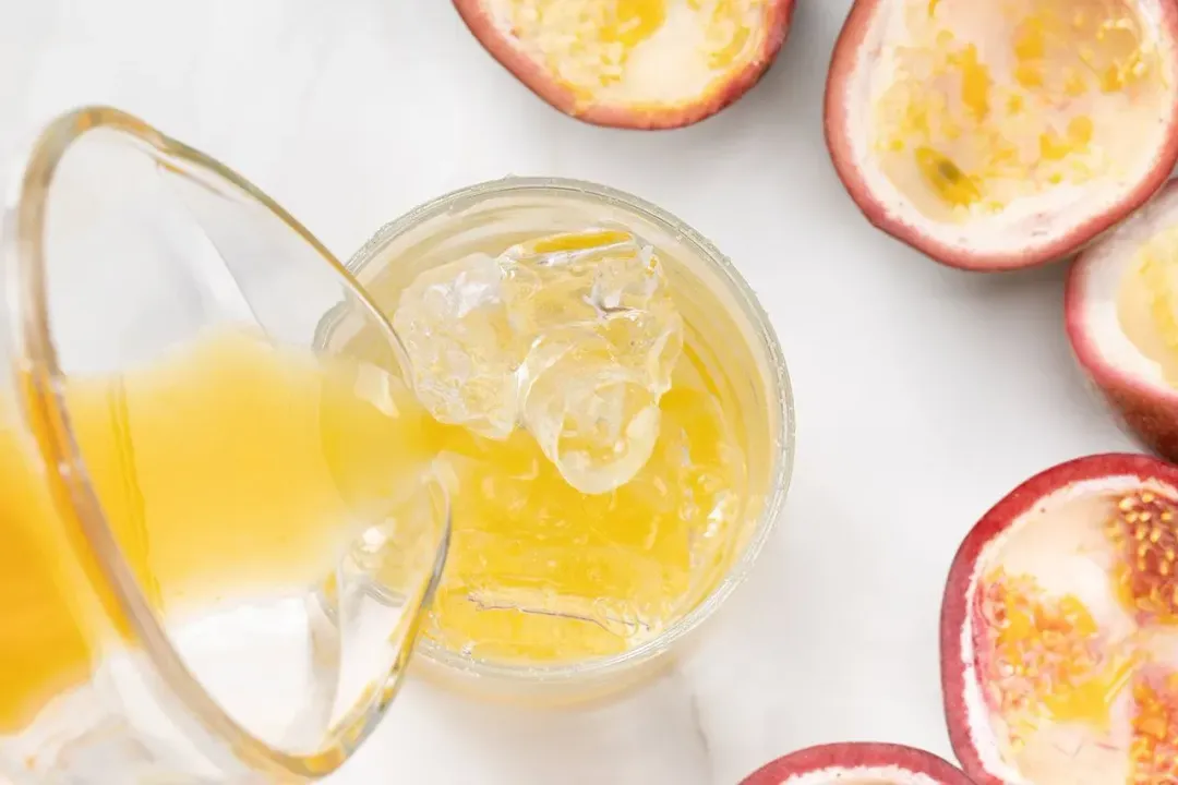 Pouring a passion fruit margarita cocktail into a short iced glass, next to hollow passion fruit shells