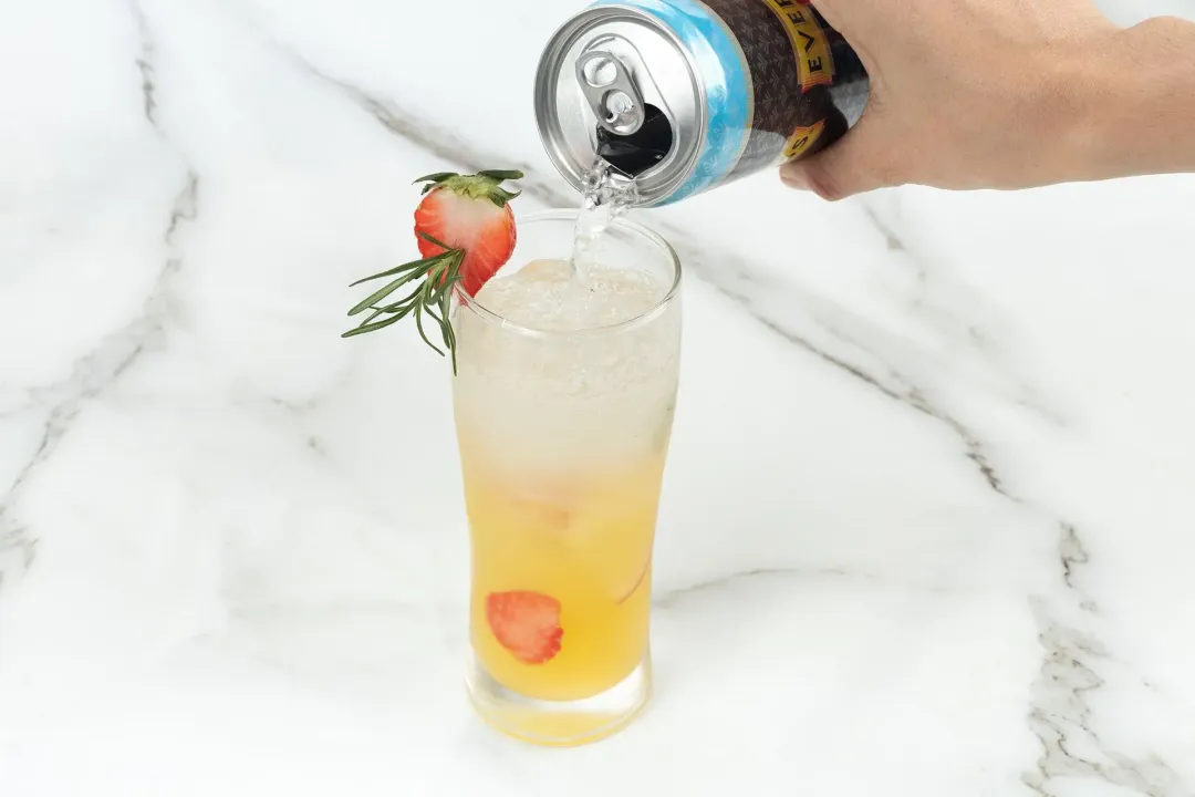 A popped soda can pouring onto a glass of orange juice vodka with ice and fruits