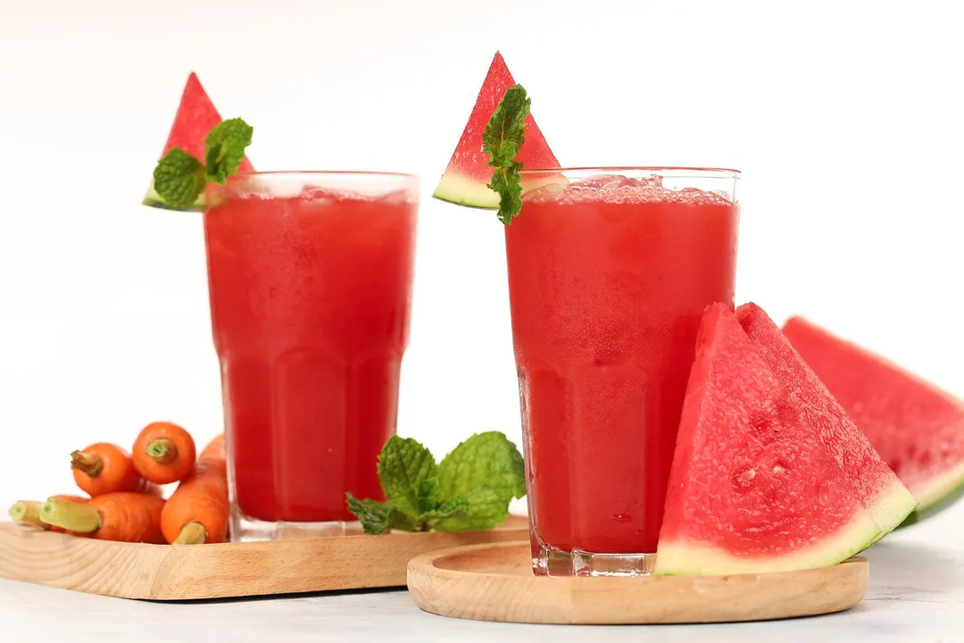 Two glasses of watermelon carrot juice placed on wooden boards near watermelon slices, mint leaves, and baby carrots.