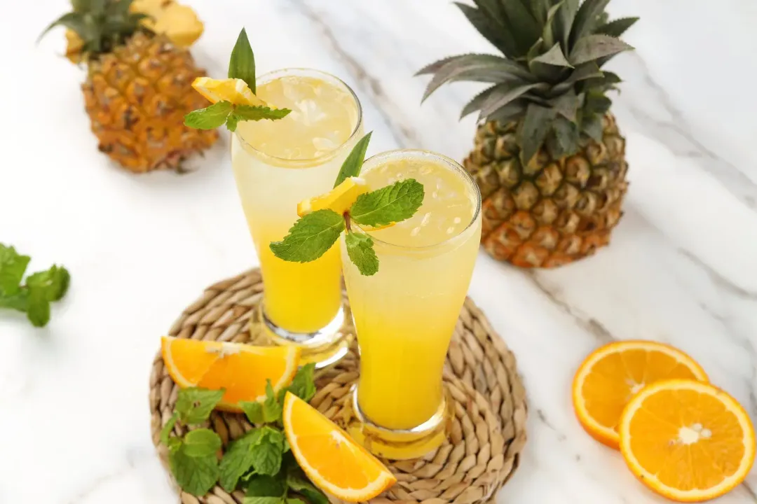 Two glasses of unstirred, iced rum and orange juice with mint sprigs, orange wedges, and pineapple tops around as garnish