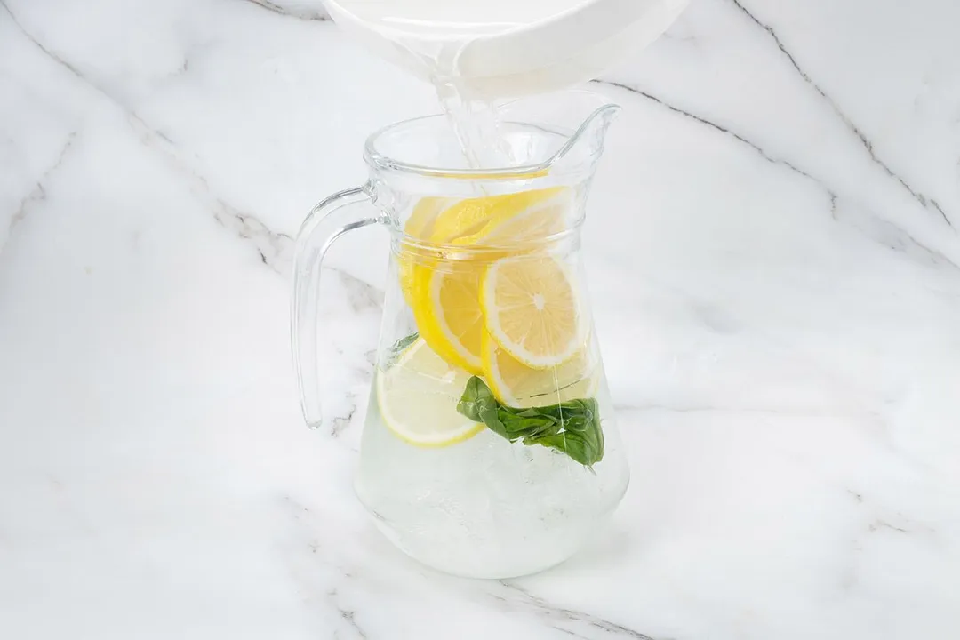 pouring water from a bowl to a jar of lemon slices and basil leaves inside it