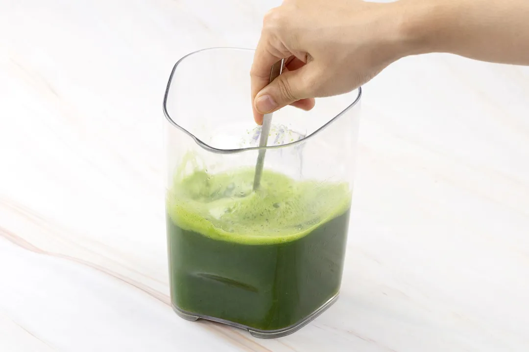 a hand holding a spoon on a pitcher of kale juice