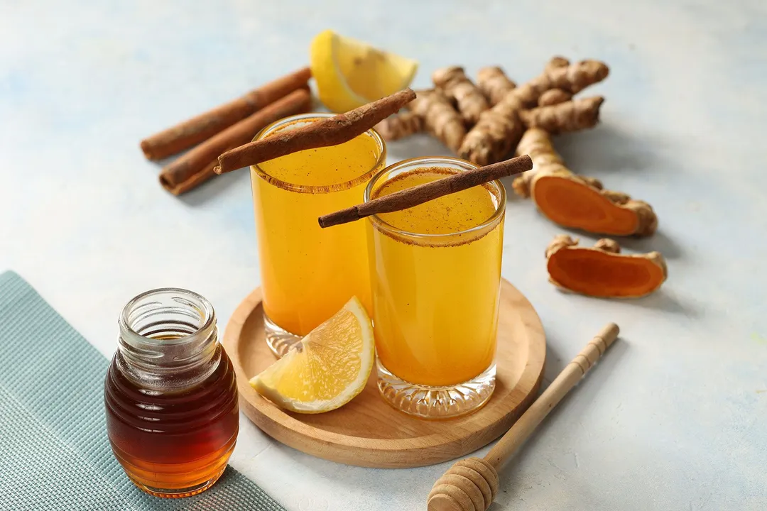 two glasses of yellow juice on a table next to a honey jar decorated with cinnamon sticks