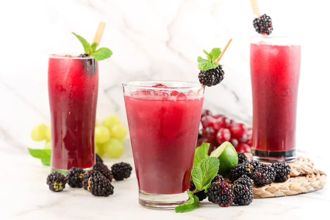Three iced glasses of black berry juice drinks with green grapes and blackberries lying around