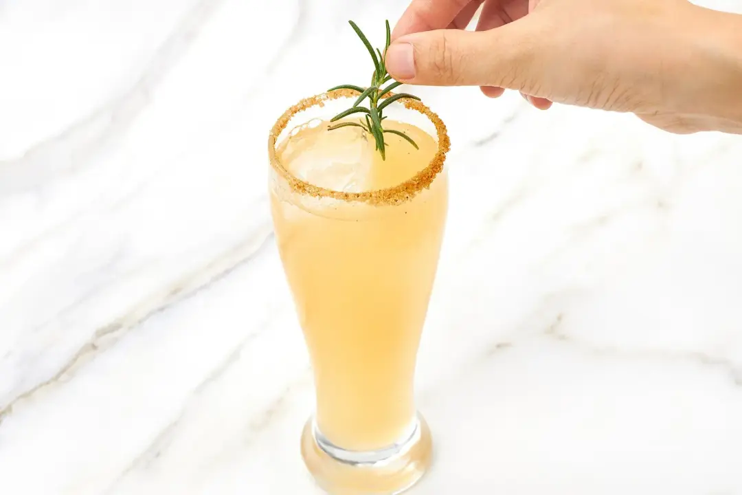 A hand putting a rosemary sprig on top of an iced apple cider margarita glass with brown sugar on the rim