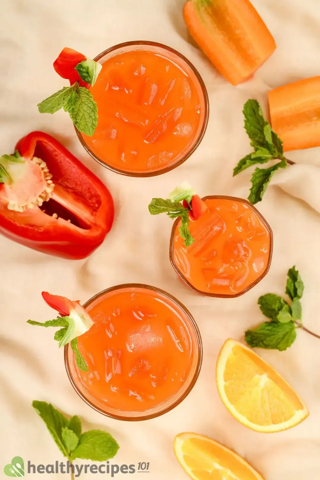 Three glasses of bell pepper juice drinks with carrots, oranges, mints, and bell pepper on a beige cloth