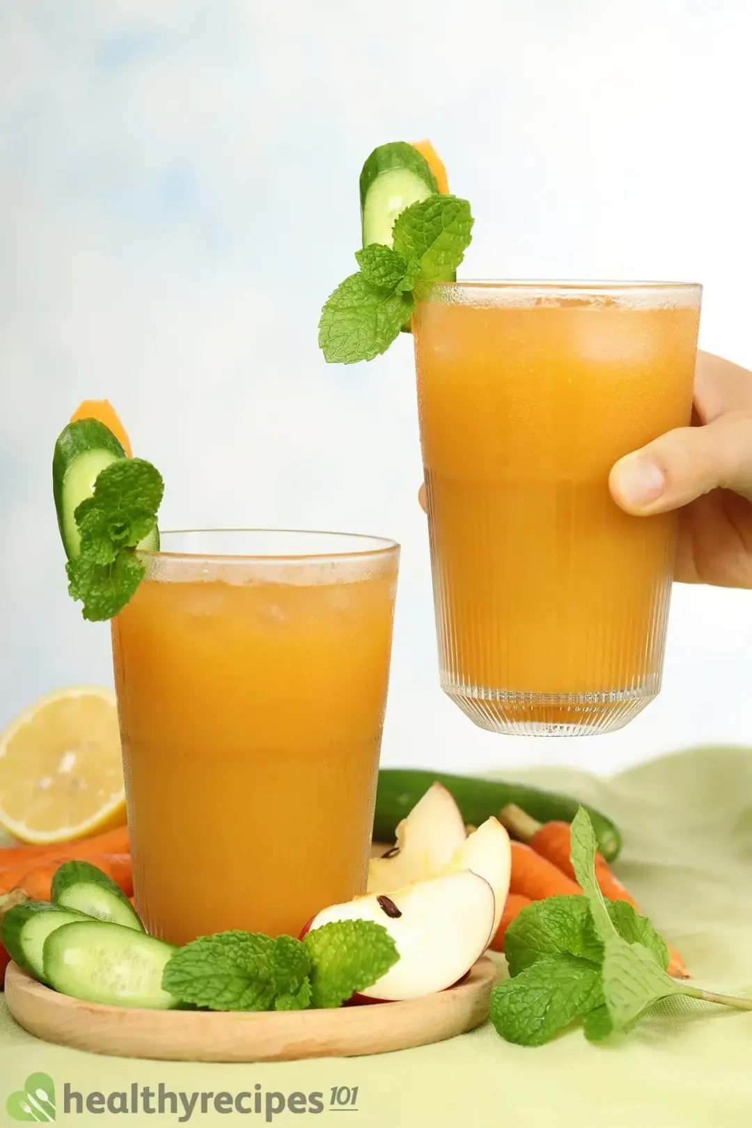 Two identical glasses of brown cucumber carrot drinks, next to sliced cucumbers, lemon wheels, baby carrots, and several apple quarters