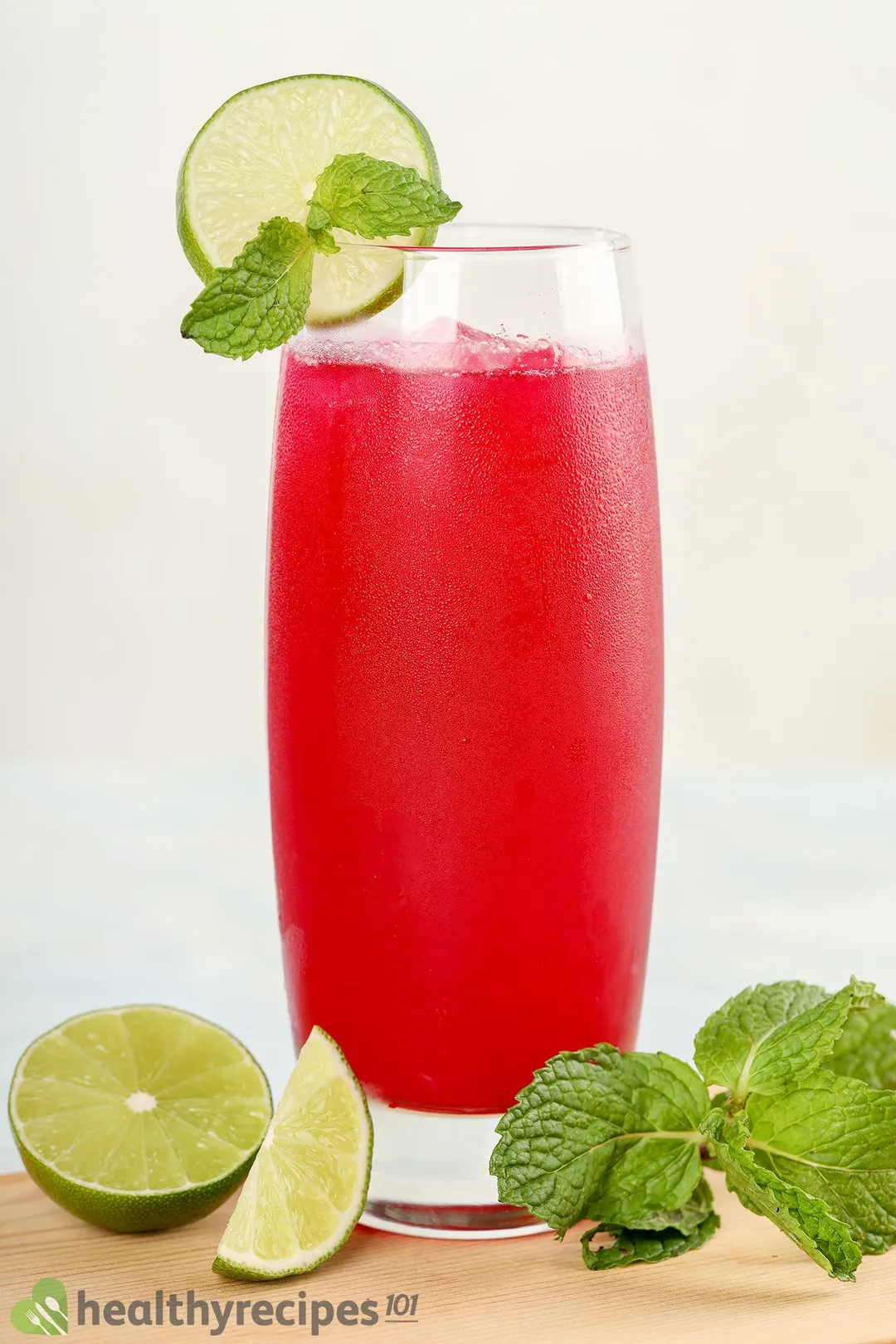 A tall, vibrant pink glass of dragon fruit juice laid near lime wedges and mint leaves. 