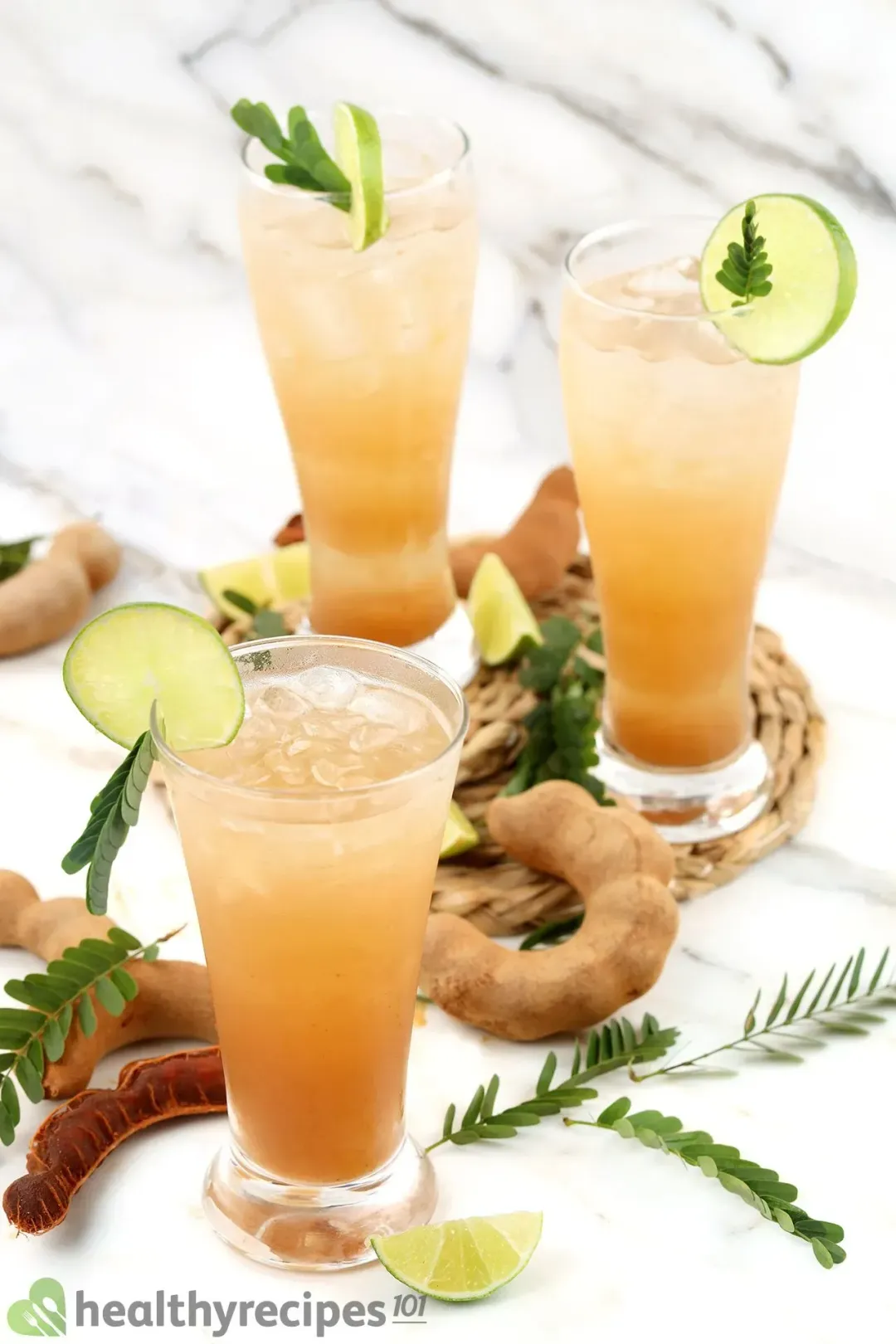 Three glasses of iced tamarind drinks with decorations: lime wheels, peeled tamarinds, and tamarind leaves