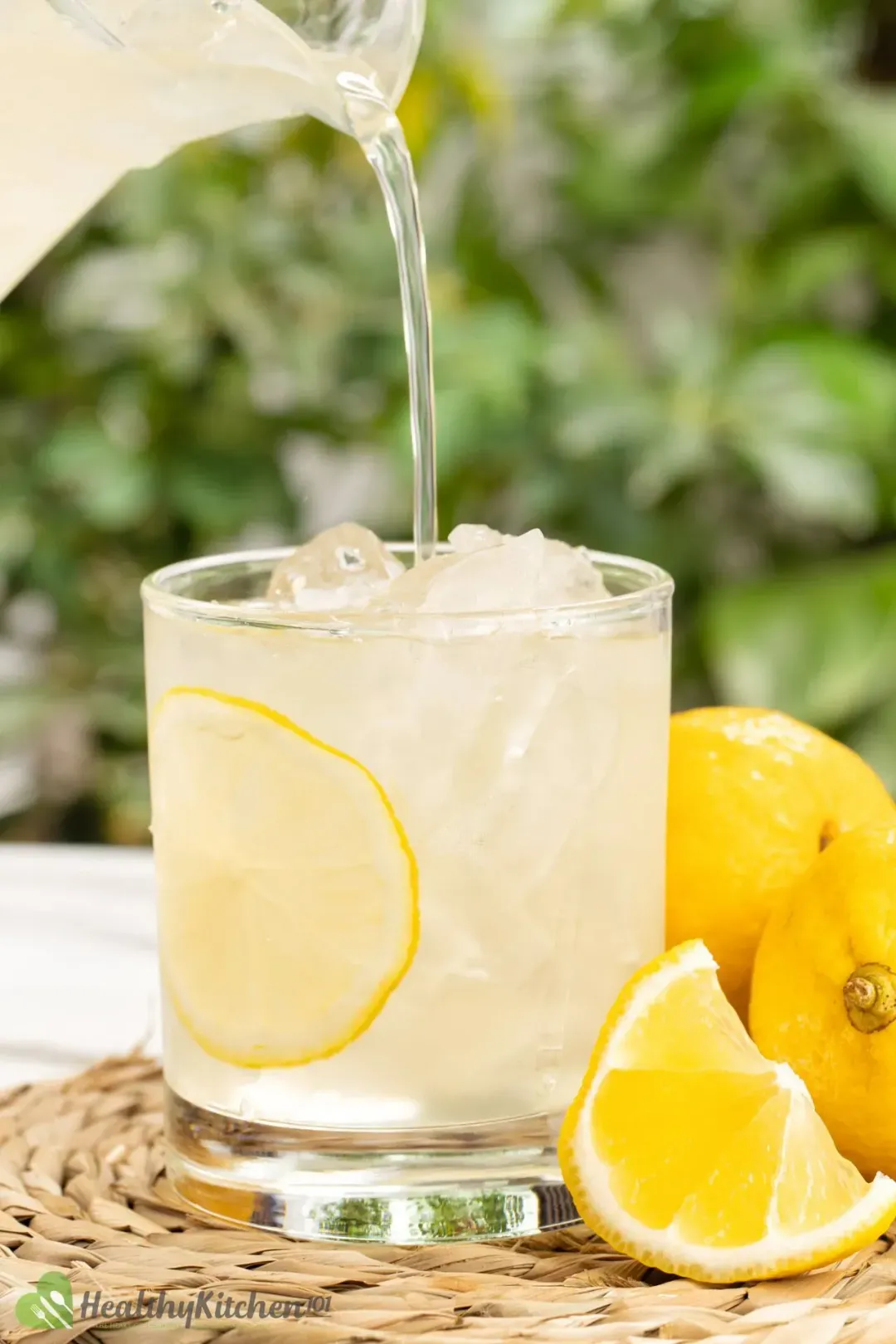 A glass of iced lemonade with lemons on the side, placed in front of green bushes