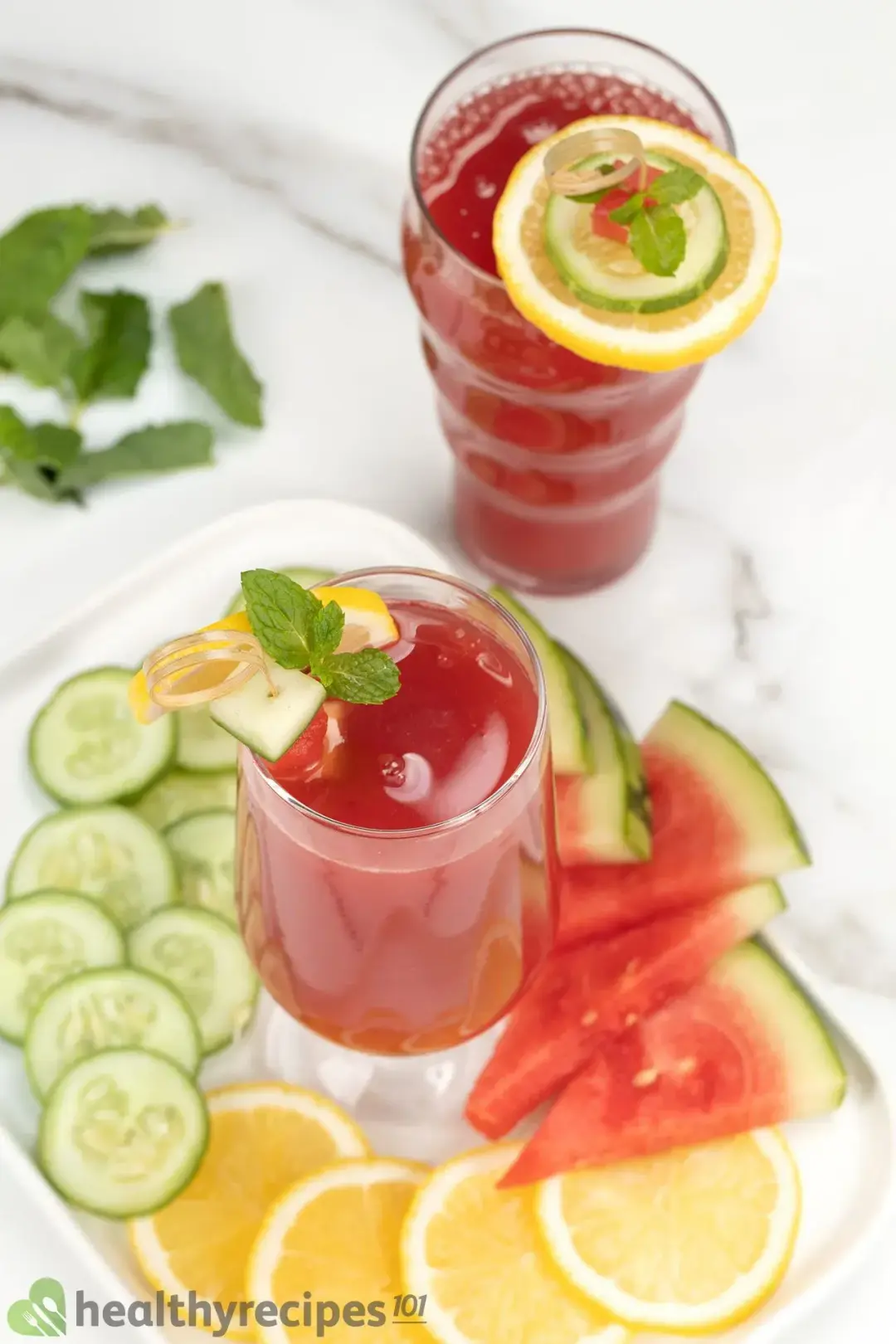 A plate holding a glass of watermelon cucumber juice surrounded by watermelon slices, lemon slices, and cucumber slices