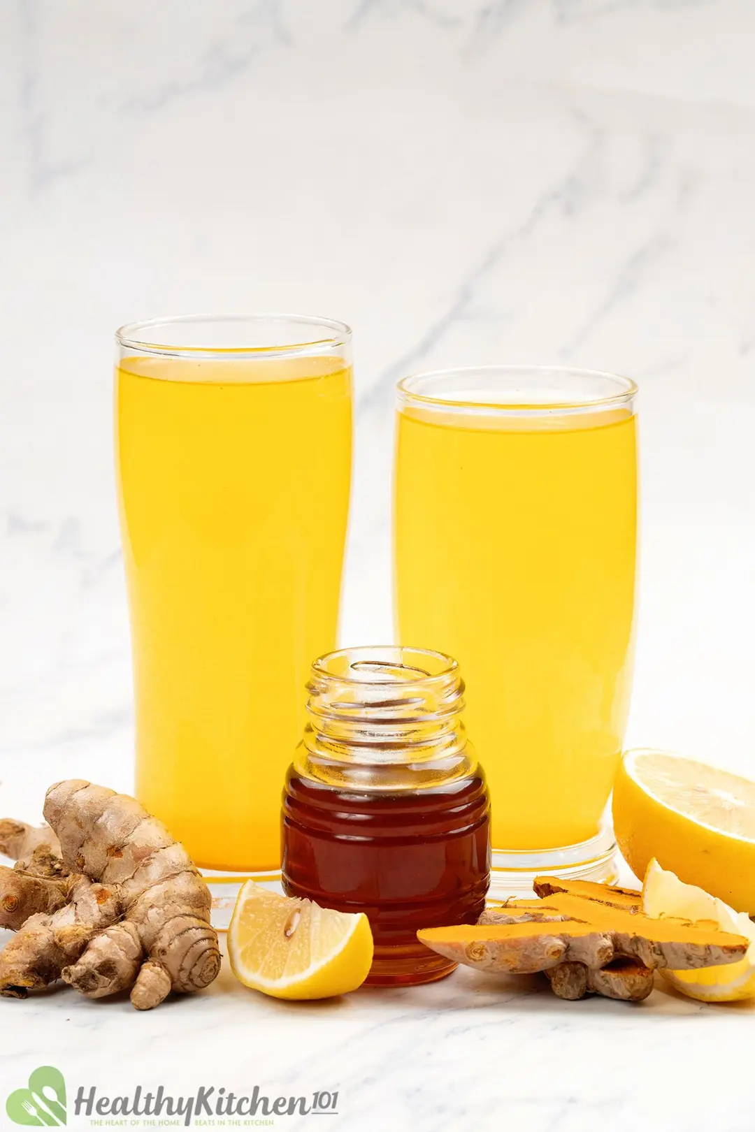 two glasses of turmeric and apple cider vinegar and a honey jar side by side, decorated with lemon and turmeric roots